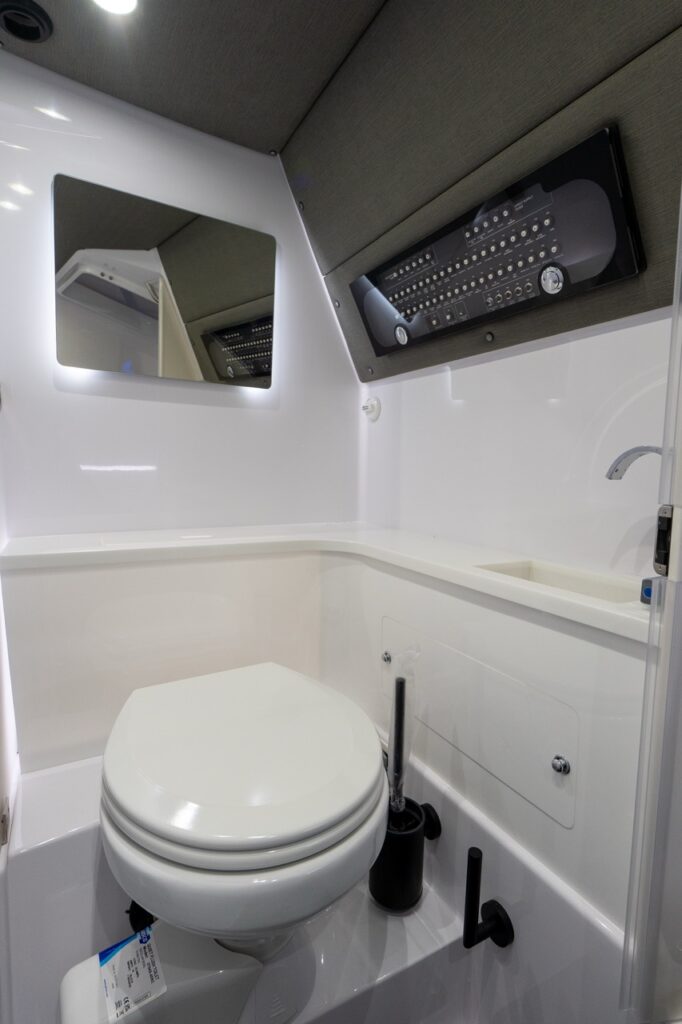 Compact enclosed marine head with a toilet in the foreground, a molded corner countertop with a small sink and faucet to starboard, and a backlit mirror on the port wall. Above the sink, a recessed black electrical switch panel is mounted under a gray upholstered liner, with bright white gelcoat surfaces and integrated lighting throughout.