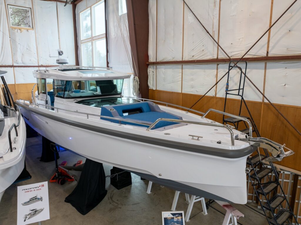 White Axopar 37 XC Cross Cabin displayed indoors on stands, viewed from the bow with blue bow sun pad cushions, enclosed cabin, and stainless railings in a boathouse.