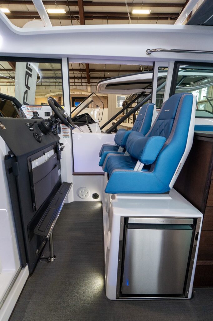 Axopar 37 XC pilot house helm with two Baltic blue suspension seats beside the helm console and an integrated stainless fridge below.