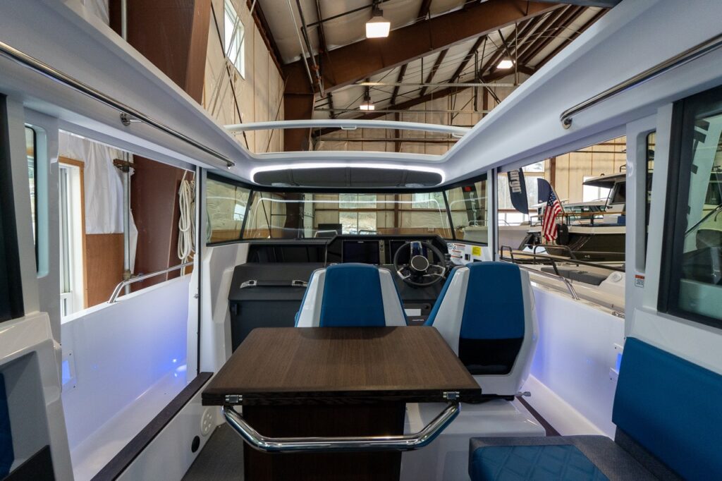 Interior view of the Axopar 37 XC Cross Cabin pilot house with two Baltic blue seats behind a wood table, looking forward to the helm and windshield.