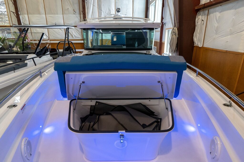 Foredeck storage compartment opened beneath the sunbed cushion on an Axopar 37 XC Cross Cabin, showing a large molded locker with gear inside and blue ambient deck lighting.