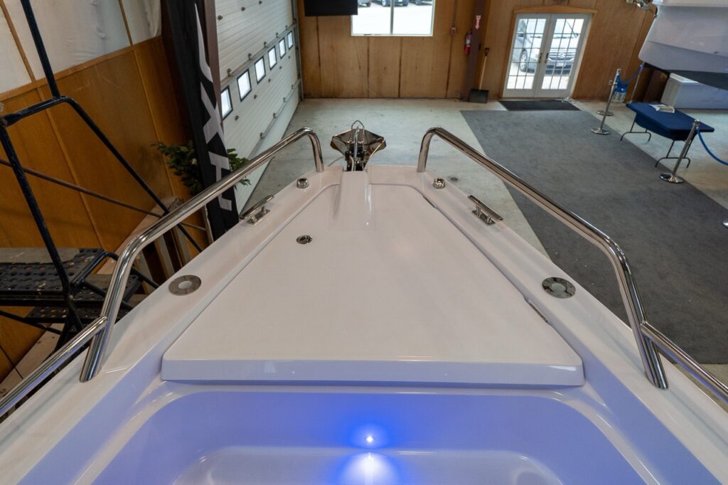 Overhead view from the roof looking down at the bow of an Axopar 37 XC Cross Cabin, showing stainless bow rails, the foredeck hatch, cleats, and anchor roller in an indoor showroom.