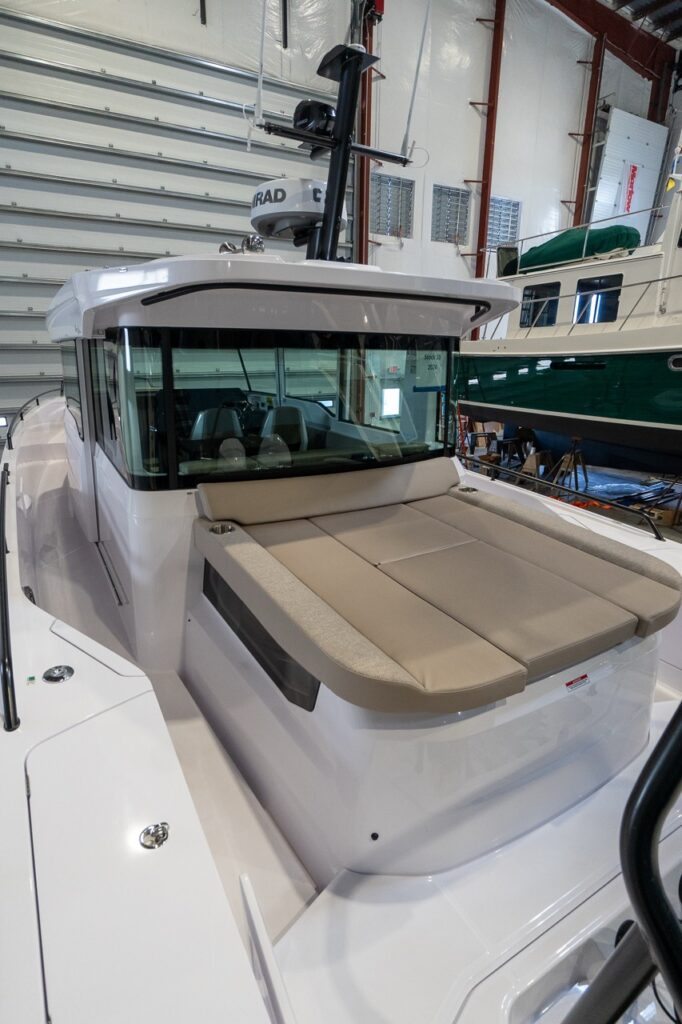 Aft deck of an Axopar 37 in an indoor marina, showing the raised sunbed with light beige cushions behind the cabin’s wraparound windows, with the roof-mounted radar mast above.