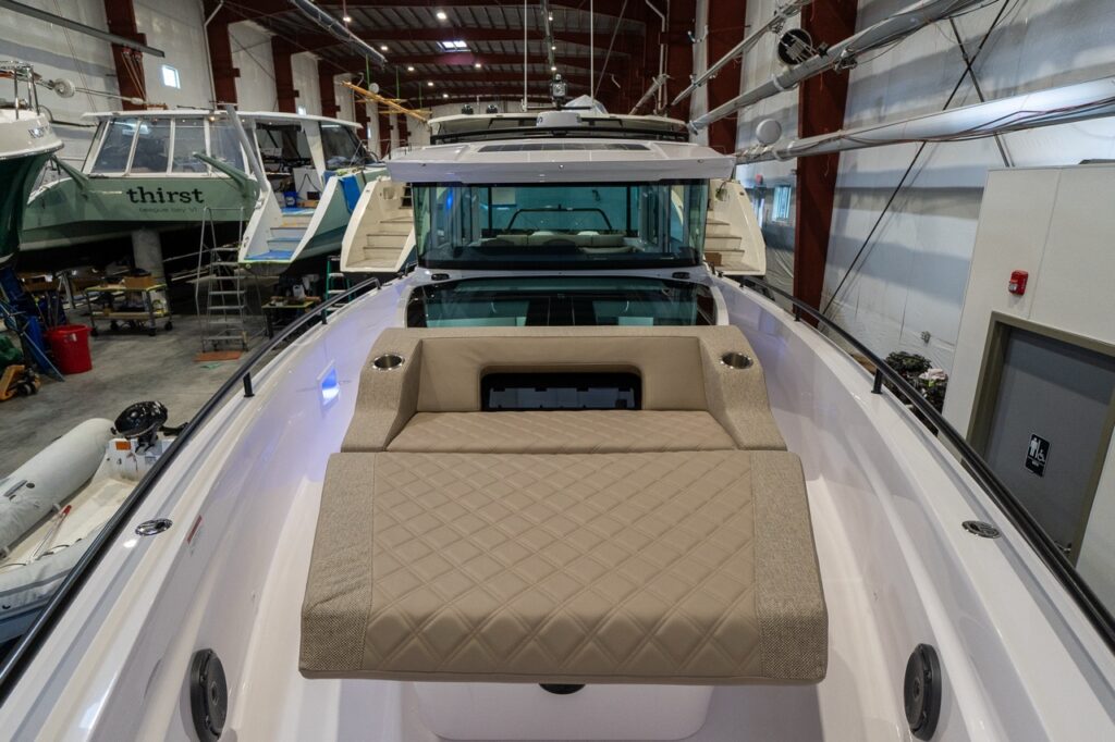 View forward along the bow of an Axopar 37 in a boat shed, featuring a large quilted Sandstone sunbed with integrated cupholders and black railings leading toward the enclosed helm and tinted windshield.