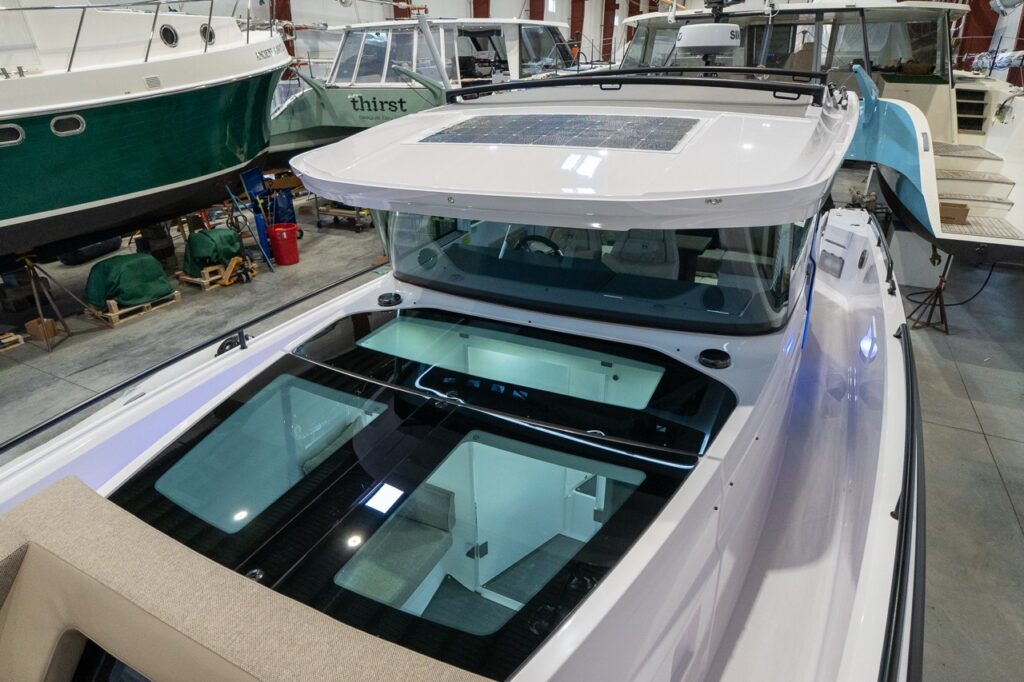 Angled view over the bow area of an Axopar 37 inside a boat shed, showing large tinted skylights in the forward cabin roof that reveal the forward cabin interior and a separate enclosed head compartment below, with the enclosed helm and roof-mounted panel visible ahead.