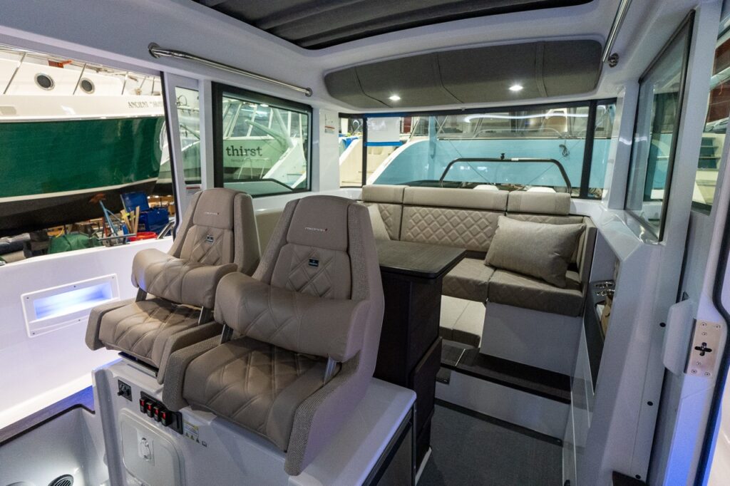 Inside the Axopar 37 wheelhouse, two Sandstone upholstered helm seats face forward beside a compact table and an L-shaped aft sofa, with large windows and overhead lighting.