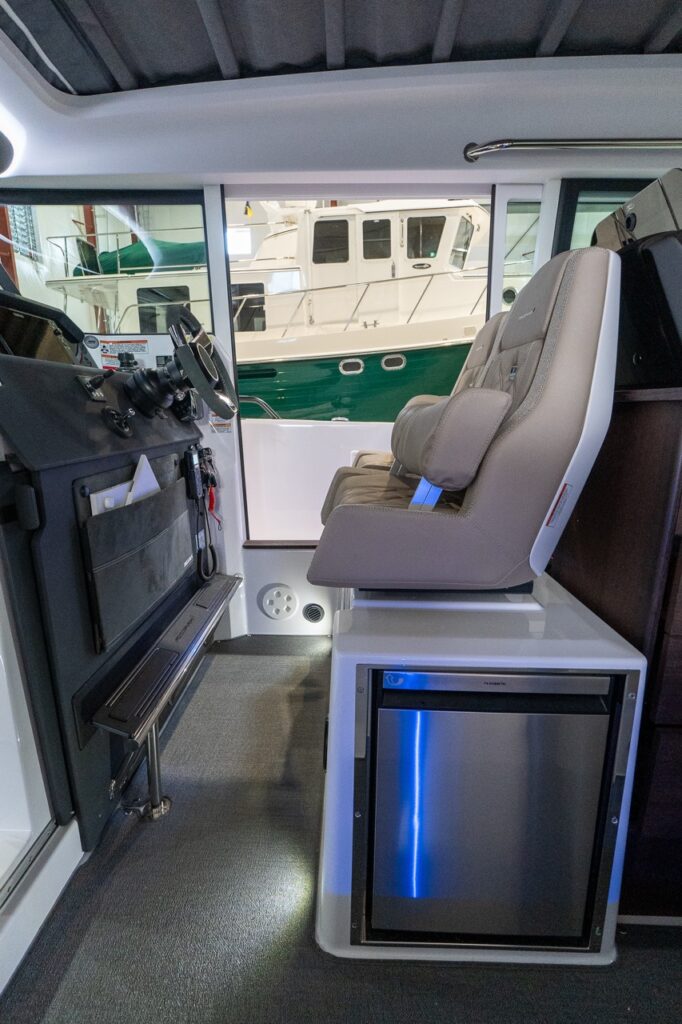 Side view inside an Axopar 37 wheelhouse showing the helm console and steering wheel opposite a Sandstone upholstered helm seat mounted above an integrated stainless-front refrigerator, with a sliding side door and marina visible outside.