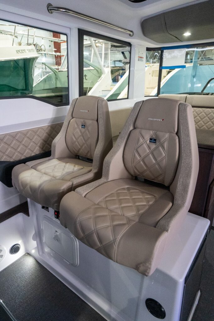 Two Sandstone upholstered Axopar 37 helm seats with diamond stitching and flip-up bolsters sit side-by-side inside the wheelhouse, framed by large windows and a quilted side panel.