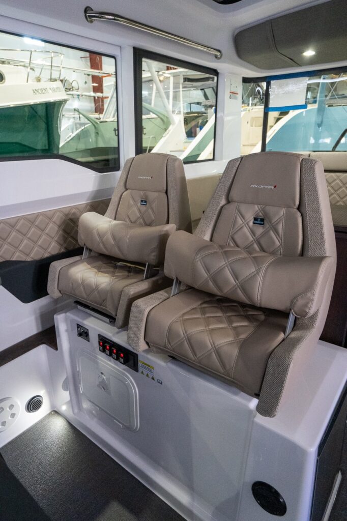 Two Sandstone Axopar 37 helm seats with diamond stitching and raised flip-up bolsters sit on a molded pedestal with integrated switches, beside quilted side padding and large cabin windows.