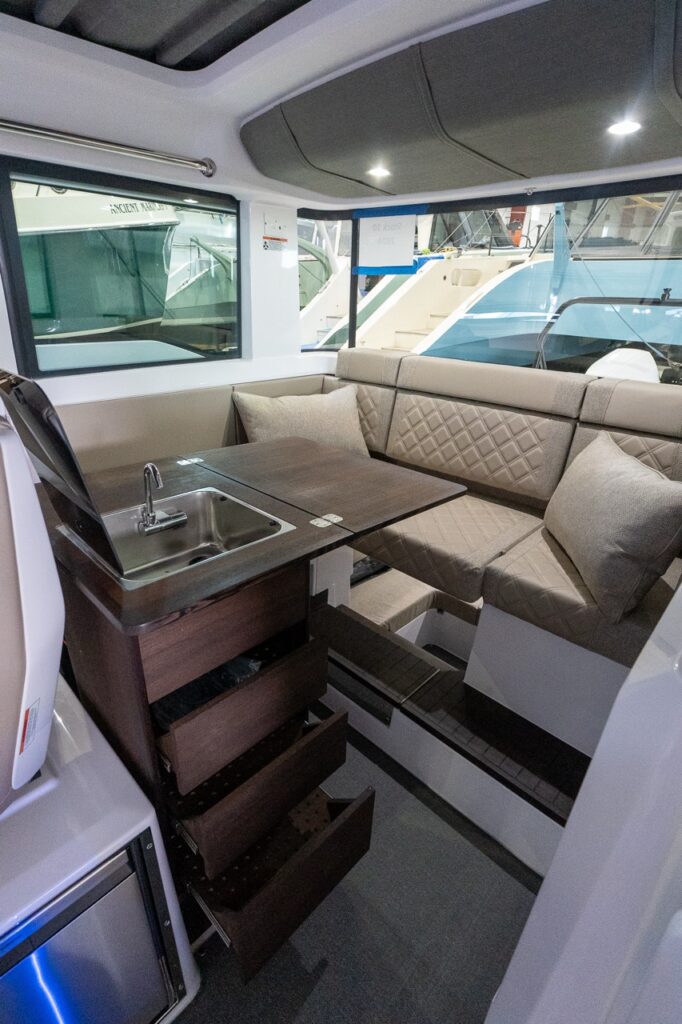 Inside an Axopar 37 cabin, a compact galley module with a stainless sink and faucet sits beside a dark wood table and L-shaped Sandstone dinette with quilted upholstery and two throw pillows.