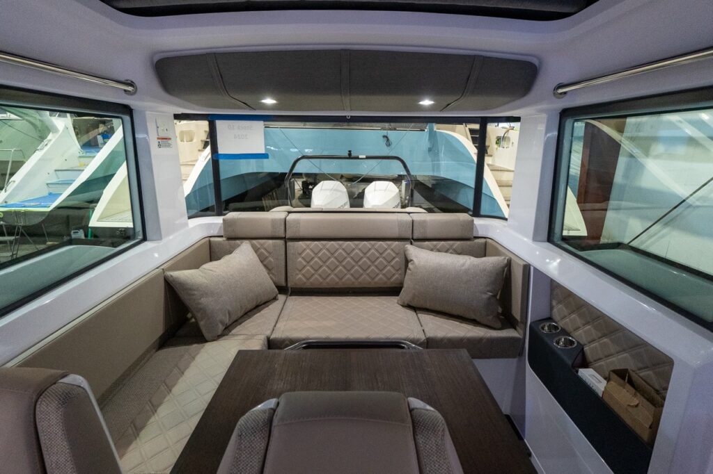 Forward-facing view inside an Axopar 37 cabin showing an L-shaped Sandstone-upholstered lounge with diamond stitching, two throw pillows, a dark wood table in the foreground, and wide side windows.