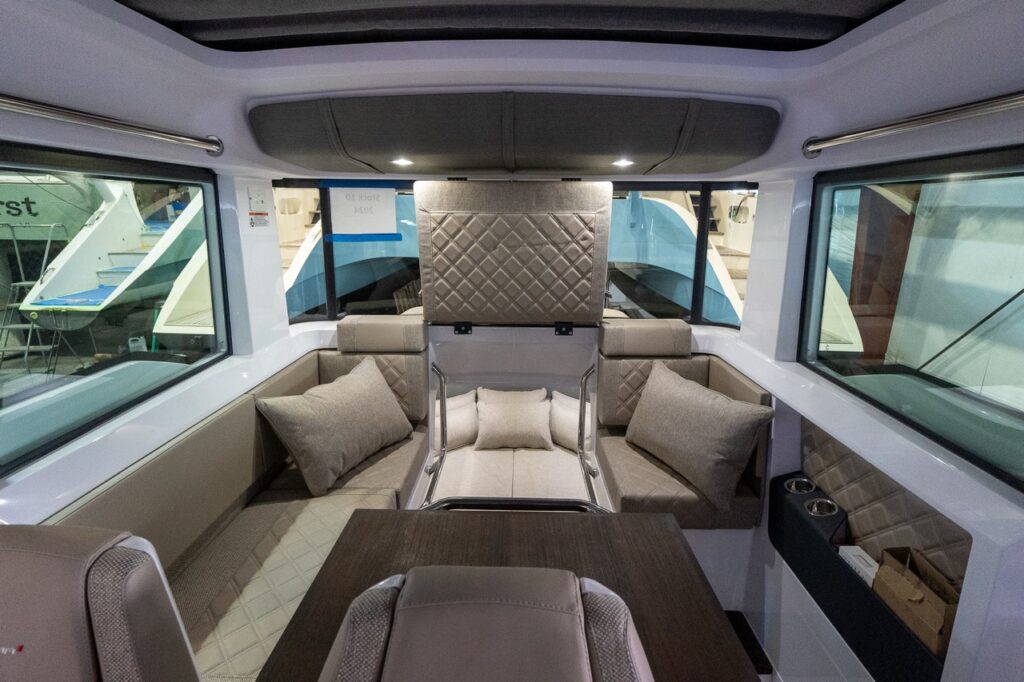 Inside the Axopar 37 pilot house, the center section of the aft bench is flipped up, revealing a walk-through opening with grab rails leading down into the aft cabin; Sandstone quilted upholstery, pillows, and a dark wood table fill the foreground.