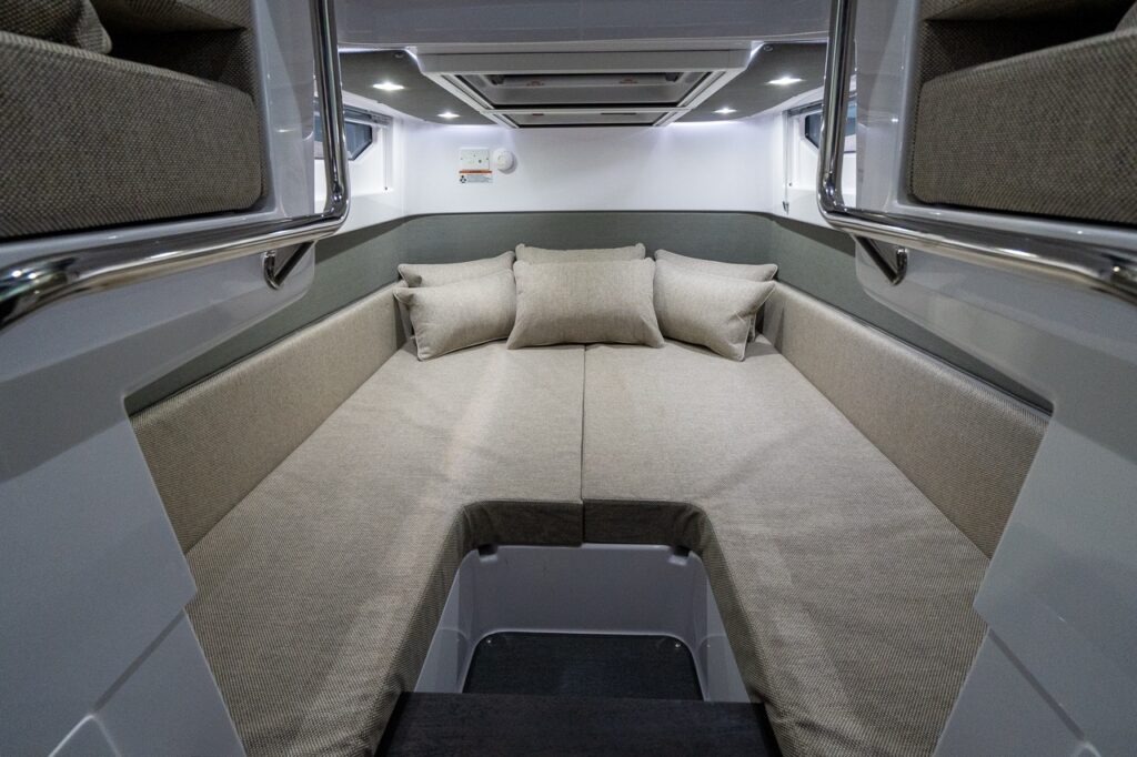 View into the Axopar 37 aft cabin showing a wide, cushioned double berth with Sandstone upholstery and three pillows, enclosed by padded side bolsters, overhead lighting, and small side windows.