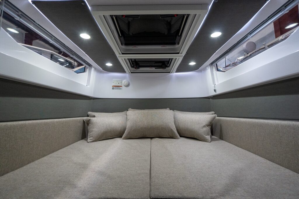 Close-up view of the Axopar 37 aft cabin berth in Sandstone upholstery, with three pillows, slim side windows, overhead hatches, and recessed ceiling lights with LED accent strips.