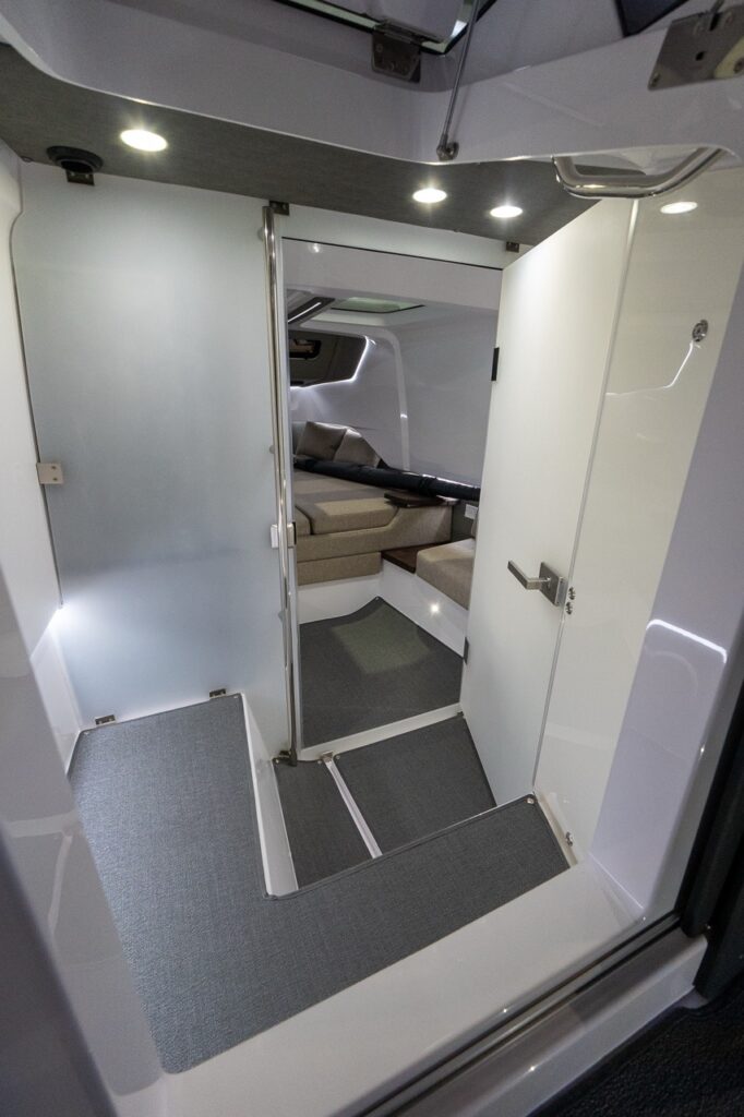 View down the steps into the Axopar 37 forward cabin, with the head compartment closed off by a door on the right and the forward berth visible ahead under soft interior lighting.