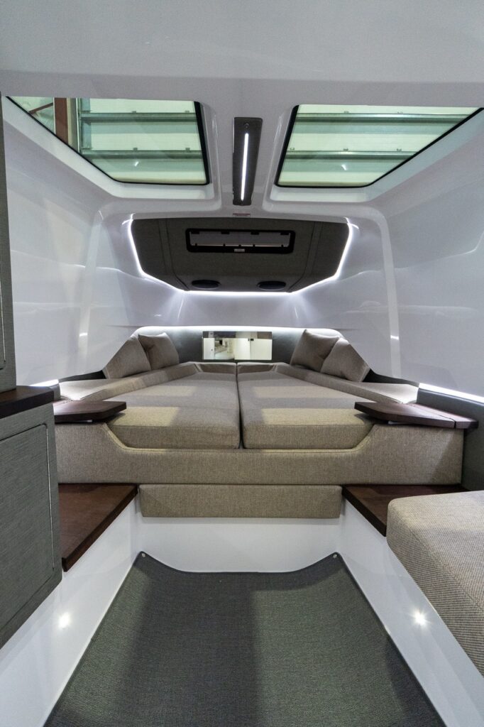 Inside the Axopar 37 forward cabin, a large V-berth/double berth with light upholstery sits beneath two overhead skylights, with ambient LED lighting, small wood side shelves, and a carpeted step in the foreground.
