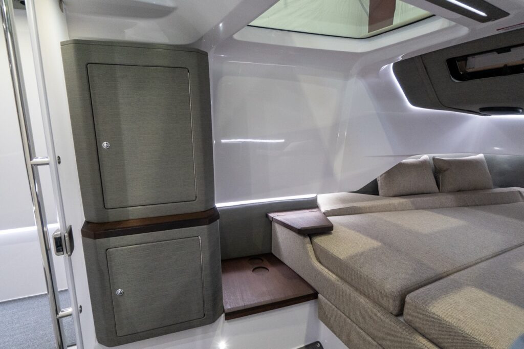 Angled view inside the Axopar 37 forward cabin showing the edge of the double berth, a wood bedside shelf, ambient LED lighting, an overhead skylight, and a tall upholstered storage cabinet with two lockable doors.