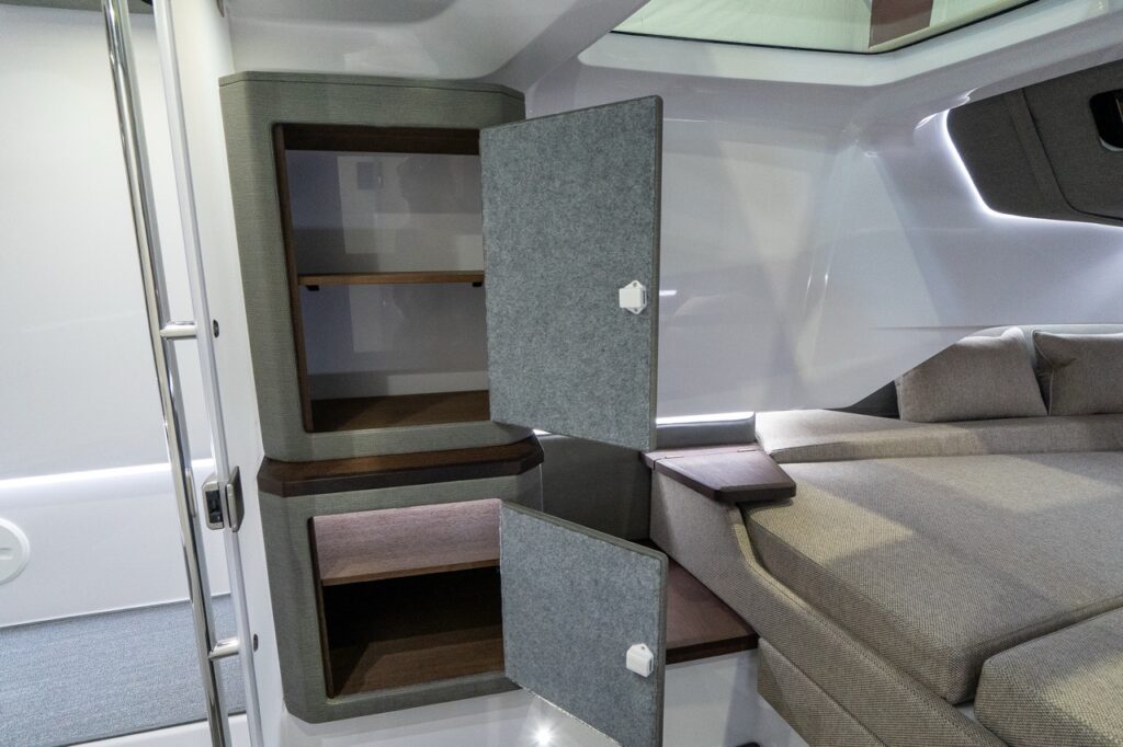 Inside the Axopar 37 forward cabin, two upholstered storage cabinets are shown with their doors open, revealing wooden shelves next to the berth and a small wood-topped bedside platform beneath a skylight and LED accent lighting.