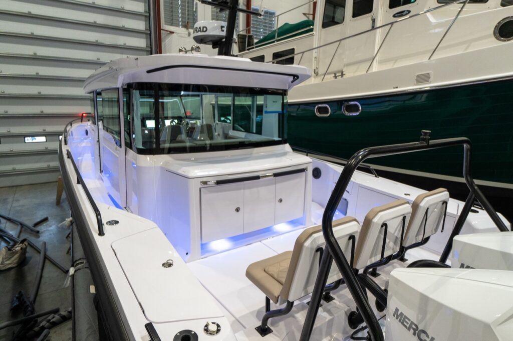 Aft deck view of an Axopar 37 XC Cross Cabin inside a warehouse, showing three fold-up “staple” jump seats in Sandstone upholstery behind the helm cabin, with twin outboards and a stainless rail frame.