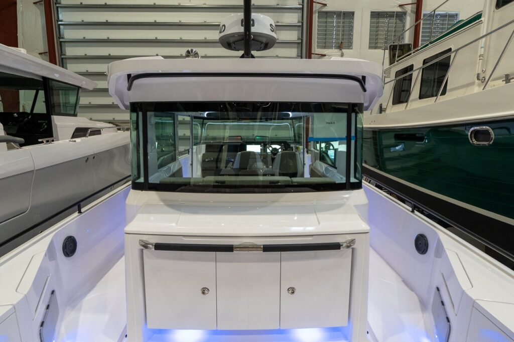 Straight-on view from the aft deck of an Axopar 37 XC Cross Cabin in an indoor storage building, showing the closed aft wet bar cabinet beneath the pilothouse rear window, flanked by wide side decks with integrated speakers and blue accent lighting.