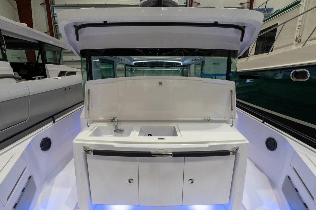 Straight-on view from the aft deck of an Axopar 37 XC Cross Cabin indoors, showing the aft wet bar with the lid raised to reveal a molded counter with a faucet, sink, and open bin, plus storage cabinets below and blue accent lighting along the side decks.