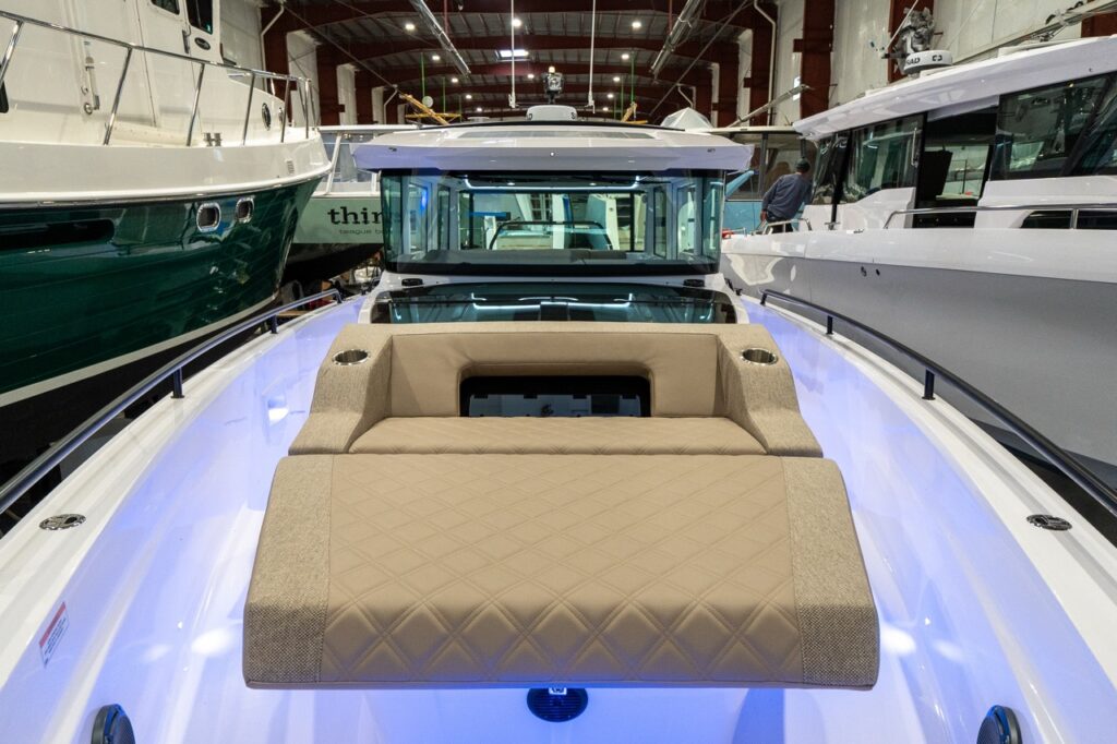 Head-on view down the foredeck of an Axopar 37 XC Cross Cabin featuring a Sandstone upholstered sunpad with diamond stitching and integrated cupholders, set between tall walkaround side rails with blue accent lighting along the deck.