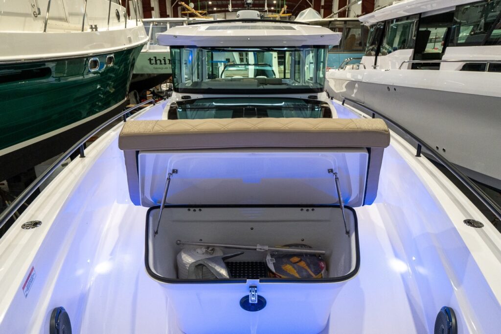 Foredeck view on an Axopar 37 XC with the Sandstone upholstered sunpad tilted up to reveal a large storage locker beneath, illuminated by blue accent lighting and surrounded by wide walkaround side decks.