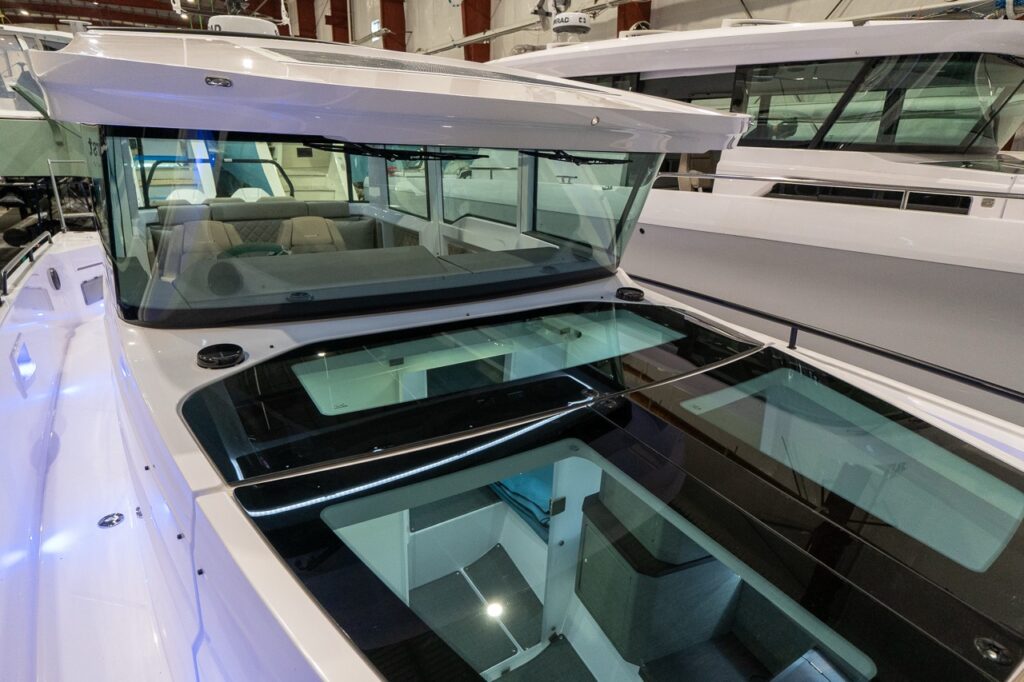 Angled view over the Axopar 37 XC’s forward cabin roof with both gullwing doors closed, forming a large tinted glass panel section; the pilothouse windshield and bright interior seating are visible through the glass.
