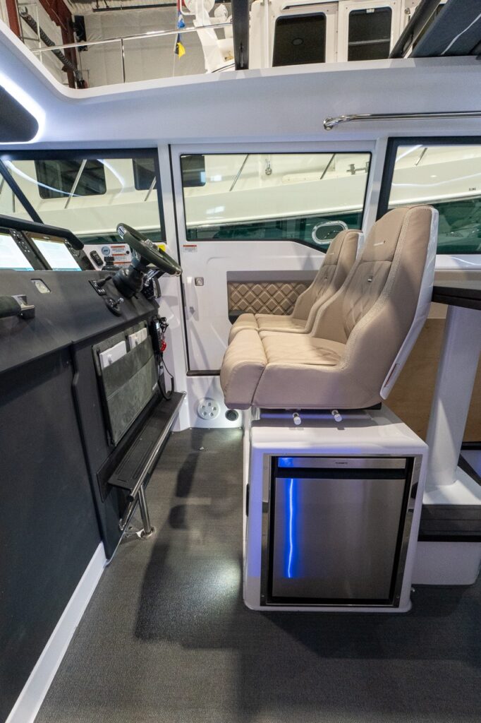 Axopar 37 XC Cross Cabin pilothouse helm area with twin helm seats, dashboard electronics, and a stainless refrigerator installed under the seats.