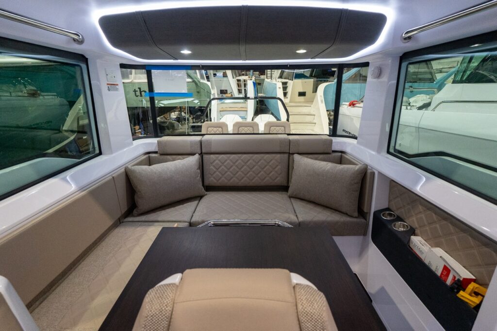 Axopar 37 XC Cross Cabin pilothouse aft lounge with wraparound seating in Dust upholstery, throw pillows, large windows, and a dining table in the foreground.