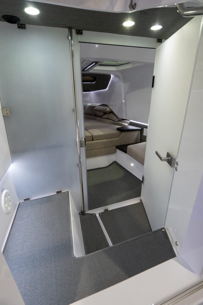 View from the Axopar 37 XC Cross Cabin enclosed head compartment looking out through the open door toward the forward berths and cabin seating, with recessed lighting and non-slip flooring.