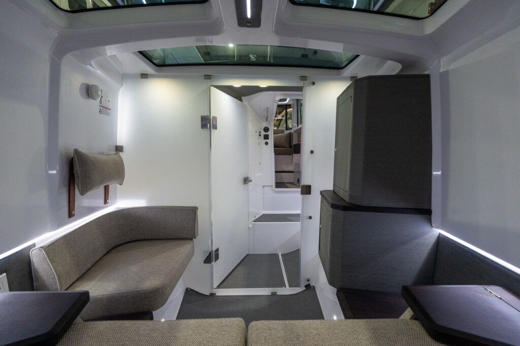 Forward cabin of an Axopar 37 XC Cross Cabin showing wraparound lounge seating, LED accent lighting, overhead skylights, and the enclosed head door open toward the companionway.