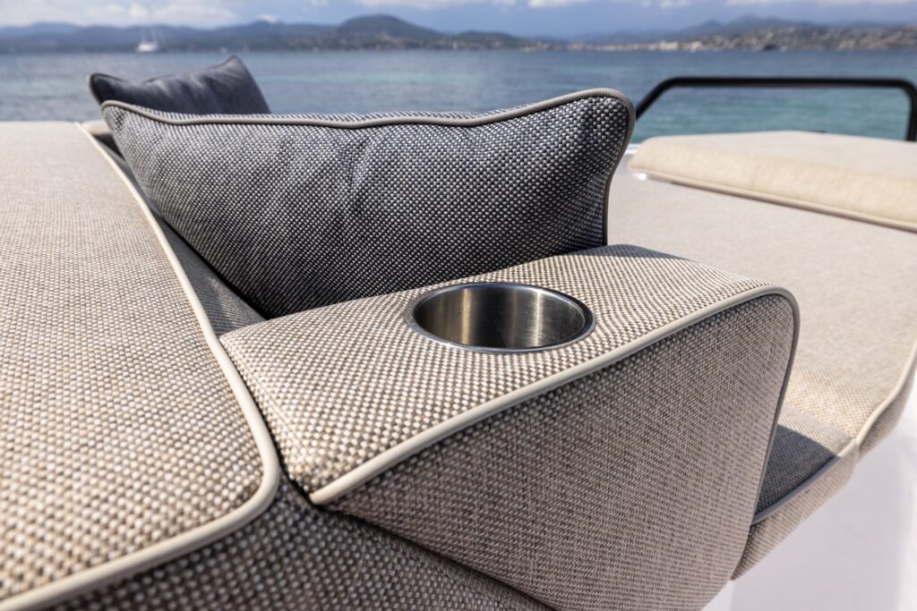 Close-up of an Axopar 37 XC foredeck sunpad showing textured upholstery, a backrest cushion, and a stainless cupholder with the sea and mountains behind.
