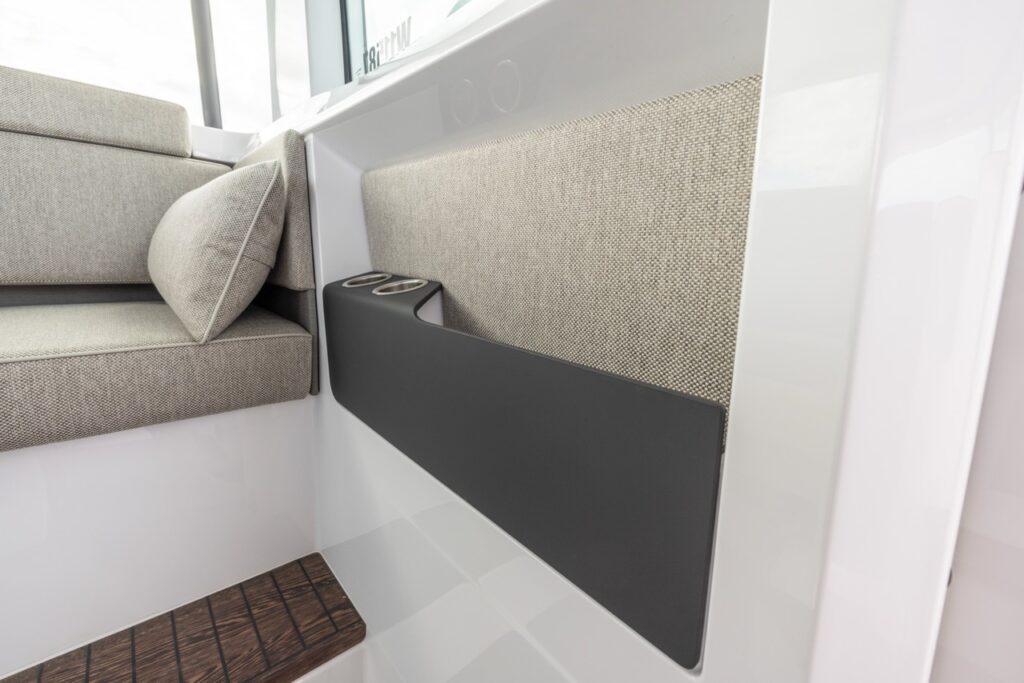 Detail view in the Axopar 37 XC Mediterrana Edition pilothouse showing textured side upholstery, corner lounge seat cushion, and integrated twin cupholders in a dark side console above teak-style flooring.