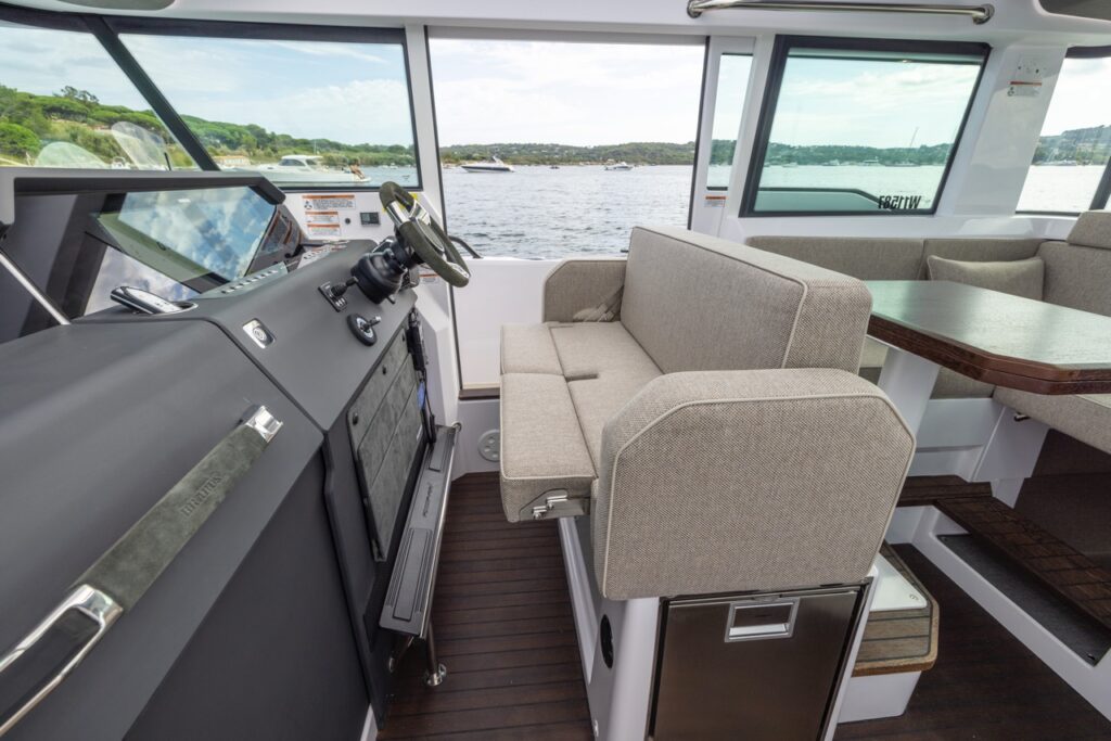 Inside the Axopar 37 XC Mediterrana Edition pilothouse showing the helm console with steering wheel and displays, a fold-down co-pilot bench seat, and aft dinette seating with a dark wood table and large windows overlooking the water.