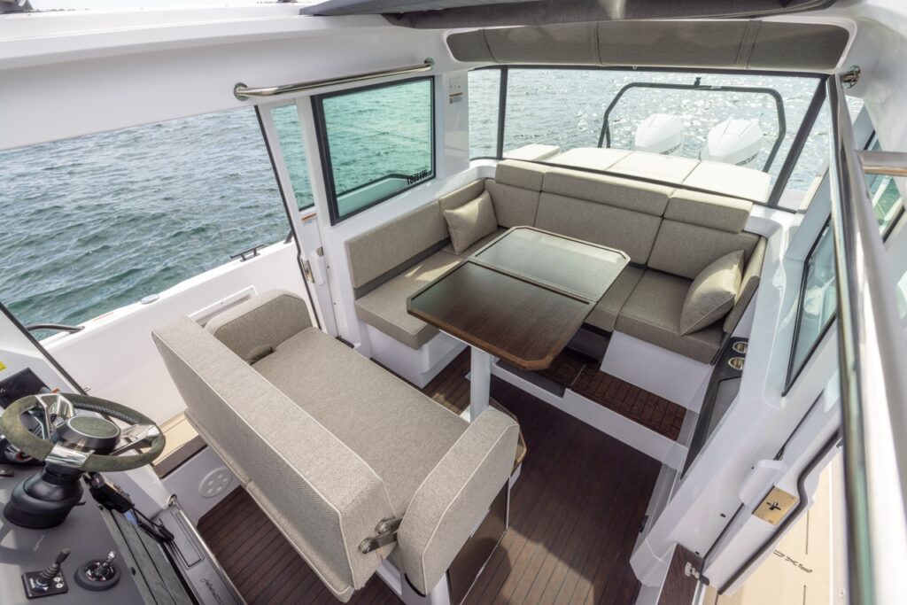 Axopar 37 XC Mediterrana Edition pilothouse with dinette table, L-shaped sofa seating, and helm seat, looking aft toward the engines.