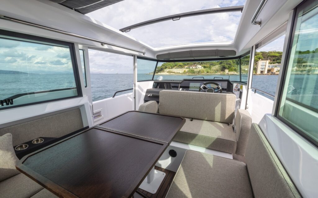 Axopar 37 XC Mediterrana Edition pilothouse dinette with large table and wraparound seating, looking forward to the helm with panoramic windows and open sunroof.