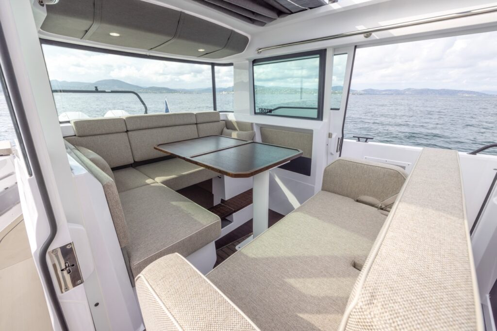 Axopar 37 XC Mediterrana Edition pilothouse dinette with U-shaped bench seating and pedestal table, surrounded by large windows overlooking the water.