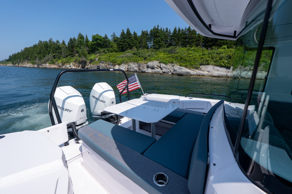 Aft seating and dining area on a 37 Axopar featuring petrol-colored cushions, a white table, and twin white Mercury outboards against a rocky shoreline.
