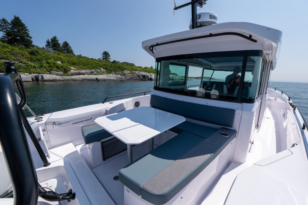 Aft dining lounge on a 37 Axopar with petrol-colored seating, a white table, and rocky shoreline in the background.