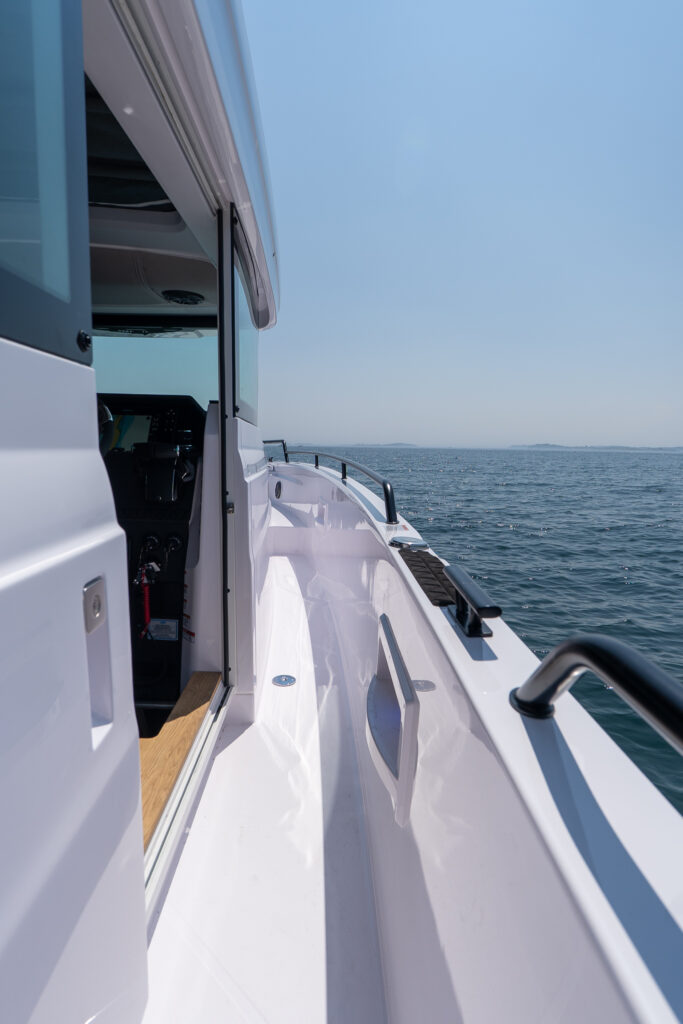 Side deck walkway of an Axopar 37 with open helm door and sturdy grab rails overlooking calm open water.