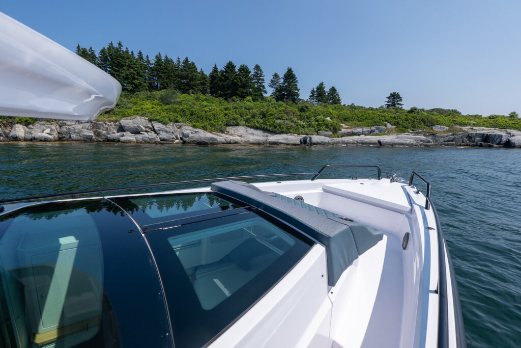 Axopar 37 XC Cross Cabin foredeck view showing the walkaround side deck, bow rails, and integrated sun pad above the cabin roof near a rocky shoreline.