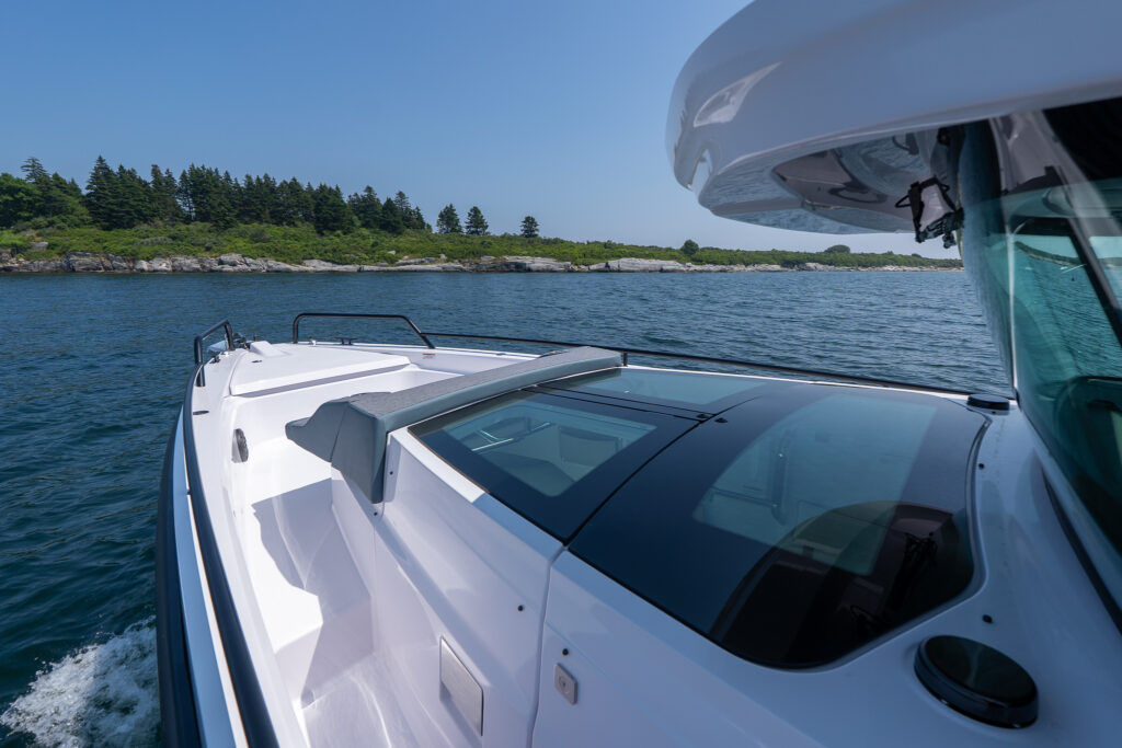 Axopar 37 foredeck featuring a diamond-stitched sunpad with integrated cupholders and gullwing doors, shown offshore with rocky shoreline in the background.