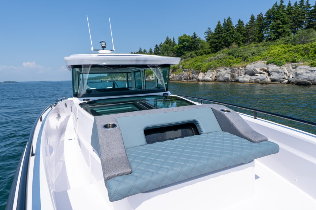 Axopar 37 XC Cross Cabin bow sun lounge on the foredeck with quilted cushions, cup holders, wraparound rails, and the pilothouse windshield in the background near a rocky shoreline.