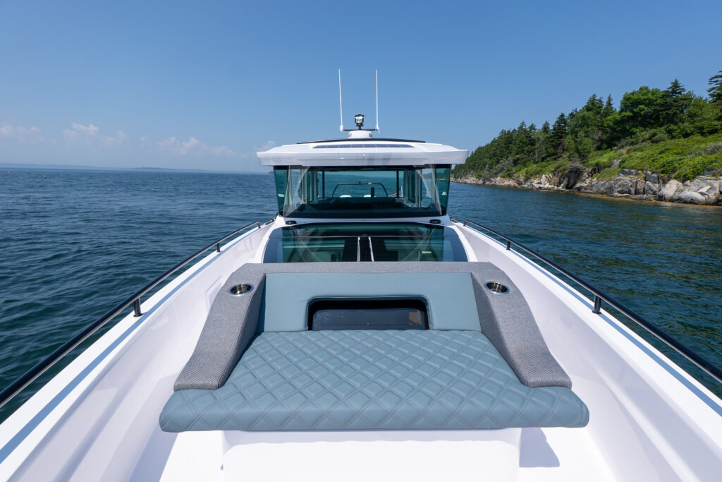 Foredeck of an Axopar 37 featuring a diamond-stitched sunpad with built-in cupholders, wide side decks, and large cabin skylights, with ocean and rocky shoreline views ahead.