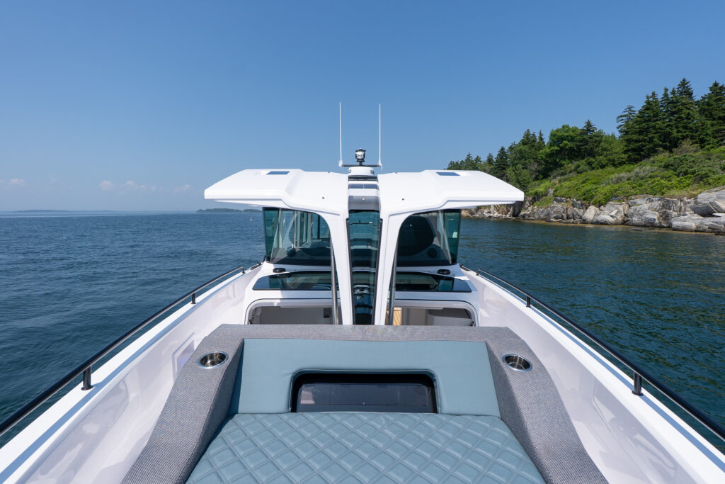 Foredeck of an Axopar 37 featuring a diamond-stitched sunpad, twin opening cabin hatches, and wide side decks, with open ocean and rocky shoreline in the background.