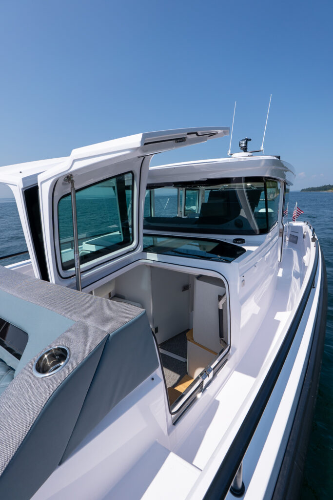 Forward cabin hatch open on an Axopar 37, revealing access to the interior berth, with cushioned sunpad seating and the wide foredeck walkway visible along the starboard side.