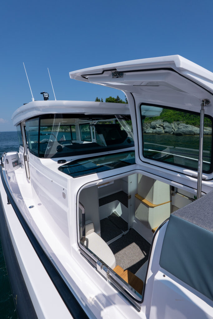 Open forward cabin hatch on an Axopar 37 showing access to the interior berth area, with large wraparound windows and the foredeck visible under clear blue skies.