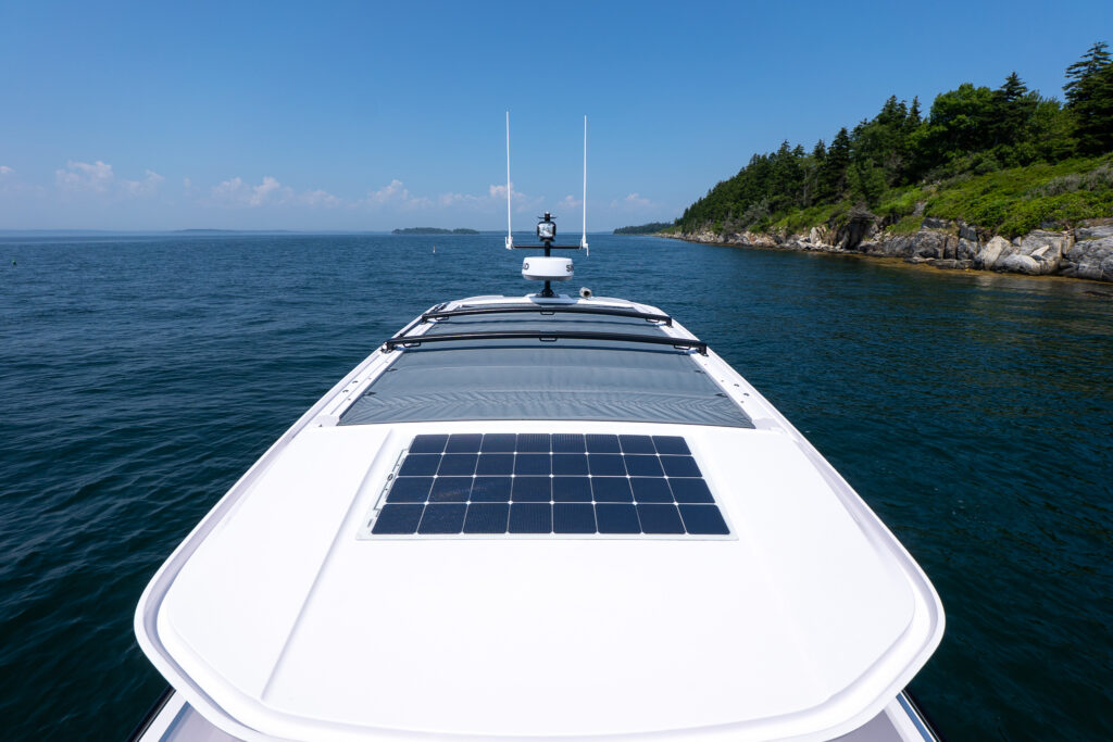 Forward view from the bow of an Axopar 37 showing the roof-mounted solar panel, sliding sunroof, Simrad radar, and dual antennae, with open ocean and a rocky, tree-lined coast ahead.