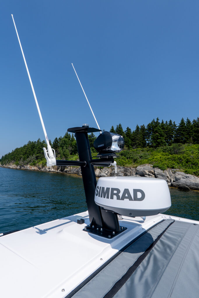 Simrad radar dome and spotlight mounted on the electronics mast of an Axopar 37, with dual antennae and a rocky, tree-lined shoreline in the background.